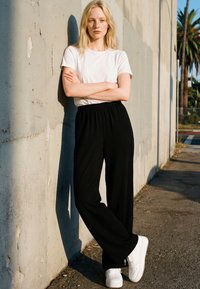 Ung kvinne med langt blondt hår står med armene i kors mot en solbelyst betongvegg, iført en hvit skjorte, svarte bukser og hvite joggesko.