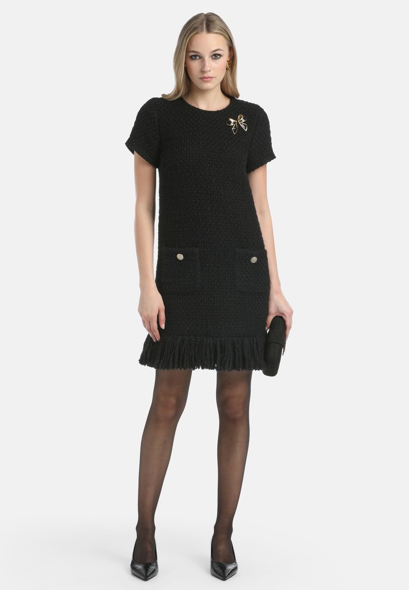 Abito nero in maglia con maniche corte, tessuto testurizzato, due tasche frontali con bottoni, dettagli con frange all'orlo, abbinato a tacchi neri.