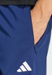 Σκούρο μπλε αθλητικό σορτς με φερμουάρ σε τσέπη και διατρητή υφή· διαθέτει λευκό λογότυπο με τρεις ρίγες στην αριστερή κάτω πλευρά.