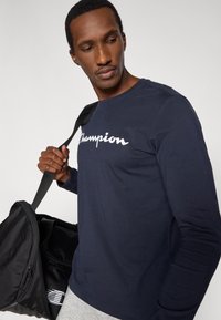 Maglietta a maniche lunghe blu navy con logo "Champion" bianco. Indossata con una borsa da viaggio nera con tasca con cerniera e pantaloni grigi.