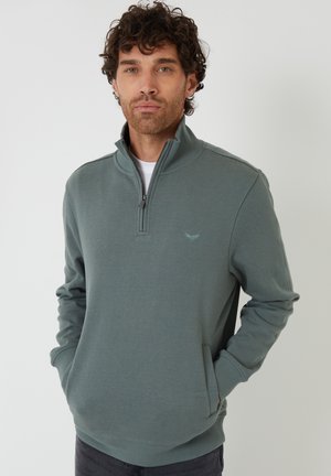 Mężczyzna z kręconymi włosami, ubrany w stonowany zielony sweter na zamek, z rękami w kieszeniach na jednolitym tle.