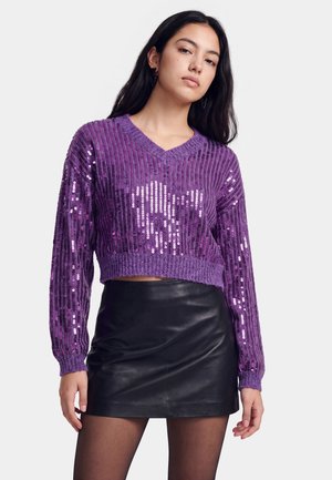 Ung kvinde iført en lilla paillet-V-hals sweater og en sort lædermini nederdel, stående mod en ensartet lys baggrund.
