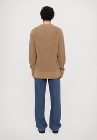 Beige strikket genser med et strukturert mønster, ribbede mansjetter og en avslappet passform, kombinert med blå vide bukser og sorte sko.