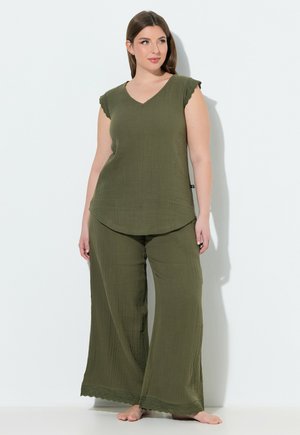 Vrouw met lang bruin haar die olijfgroene, getextureerde wijde broekspijpen en een bijpassende mouwloze top draagt, blootsvoets staand tegen een witte muur.