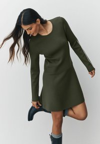 Olijfgroene jurk met lange mouwen en geribbelde textuur, met een ronde halslijn en witte zijkantstrips. Gepaard met zwarte laarzen.