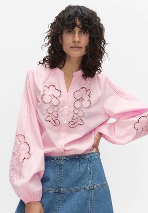 Kiharatukkainen nainen pukeutuu vaaleanpunaiseen puseroon, jossa on kukkakuvioinen leikkausommel ja liehukkeet hihansuuissa, yhdistettynä siniseen farkkuhameeseen.