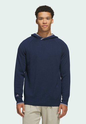 Junger Mann steht, trägt einen dunkelblauen Kapuzenpullover und beige Hosen, schaut geradeaus vor einem einfachen hellen Hintergrund.