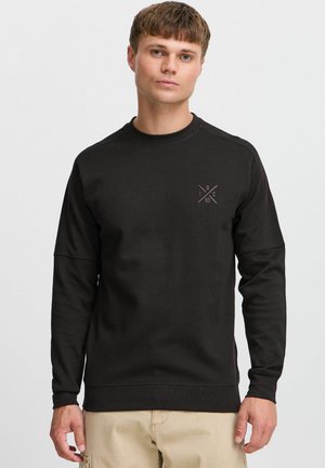 Sudadera negra hecha de tejido grueso, con cuello redondo, mangas largas y un pequeño logotipo geométrico en el pecho. Presenta costuras decorativas.