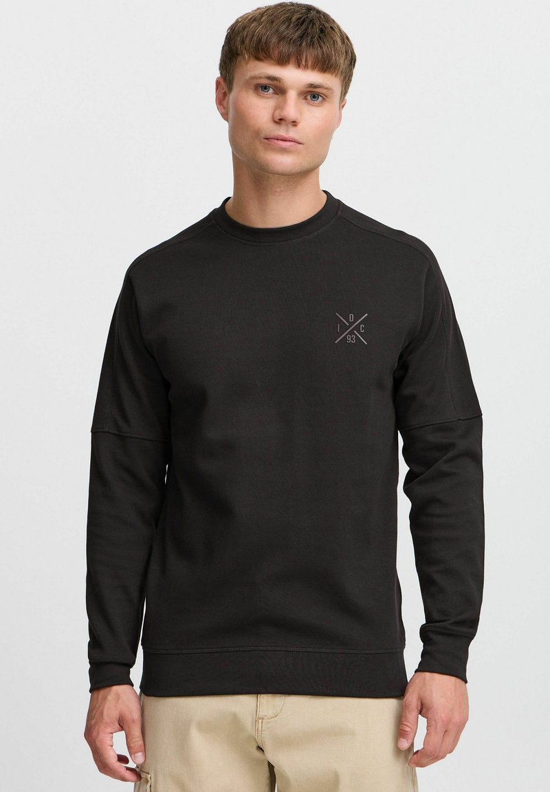 Black sweatshirt made of thick fabric, featuring a round neck, long sleeves, and a small geometric logo on the chest. Seam detailing is present.