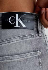 Světle šedé džíny z denimu s černou etiketou, na které je nápis "Calvin Klein Jeans" a výrazné, dominantní logo "CK" na pase.