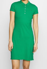 Vihreä polo-mekko, joka on valmistettu puuvillasta. Mekossa on kaulus, lyhyet hihat ja nappilista. Vasemmassa rinnassa on pieni logo.