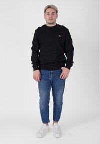 Black cotton sweatshirt with ribbed cuffs and hem, paired with blue denim jeans and white sneakers. Features a small logo on the chest.
