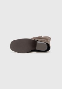 Botă din suede maroniu, de lungime medie, întinsă pe lateral, arătând o sole netedă, din cauciuc închis la culoare, cu un toc texturat, pe un fundal alb.