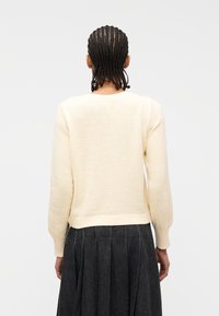 Άτομο με πλεξούδες που φορά ει κρεμ πλεκτό πουλόβερ και σκούρα πλισέ φούστα, με την πλάτη γυρισμένη, πάνω σε απλό λευκό φόντο.