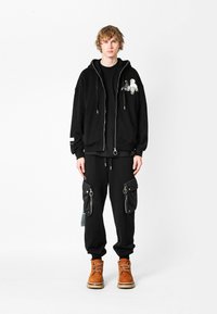 Joven de pie, vistiendo una sudadera con cremallera negra con gráfico, pantalones cargo negros con grandes bolsillos con cremallera y botas de cordones color arena.