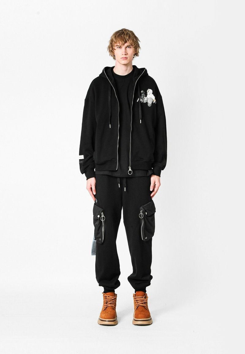 Joven de pie, vistiendo una sudadera con cremallera negra con gráfico, pantalones cargo negros con grandes bolsillos con cremallera y botas de cordones color arena.