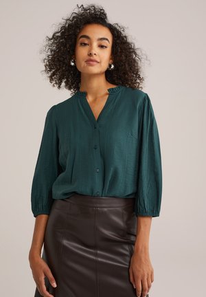 Camicia blu verde acqua con colletto plissettato, tessuto strutturato e maniche a tre quarti. Indossata con una gonna in pelle marrone aderente.