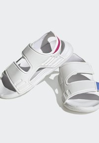 Sandali bianchi con doppie cinghie in velcro, design curvo, punta aperta, soletta texturizzata e piccoli dettagli colorati in rosa e blu.