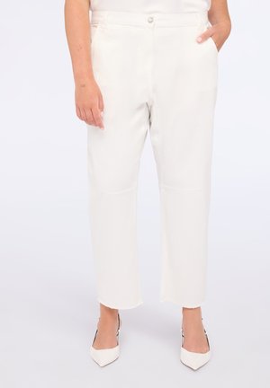 Vrouw in een hoog geproduceerde, off-white cropped broek met raw hems en witte puntige hakken, met één hand in de zak en de andere ontspannen aan de zijde.