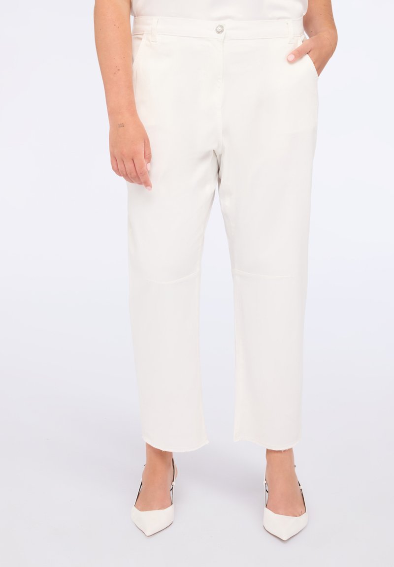 Mujer que lleva pantalones cortos de tiro alto en color blanco roto con dobladillos sin rematar y tacones blancos de punta. Tiene una mano en el bolsillo y la otra relajada a un lado.