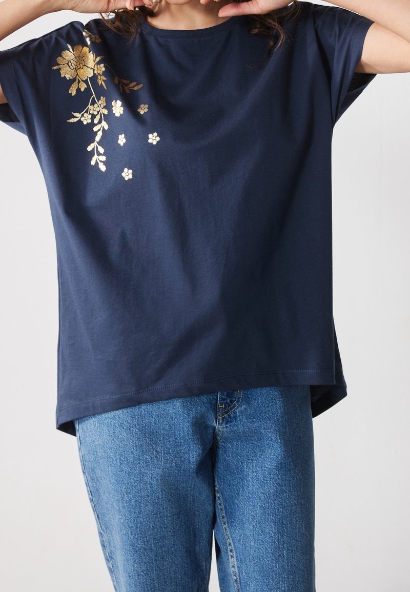 Personne portant une chemise bleu marine à manches courtes avec un motif floral doré sur l'épaule et un jean bleu, les mains près du visage.