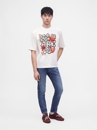 Jeune homme debout portant un t-shirt blanc avec un motif graphique, un jean bleu et des baskets rouges sur un fond clair uni.