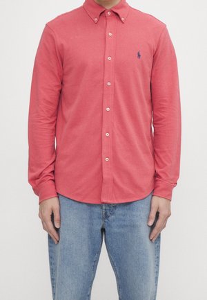 Homme portant une chemise corail à manches longues avec des boutons, avec un petit logo brodé en bleu marine sur la poitrine gauche, et un jean bleu clair.