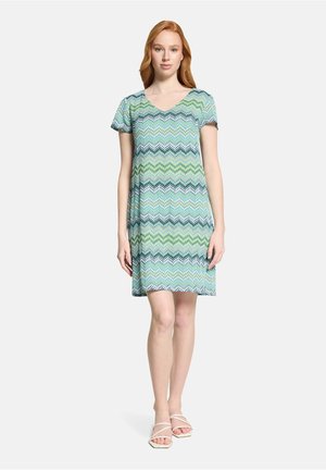 Vrouw staand, gekleed in een kort mouw, knie-lange jurk met een zigzagpatroon in blauw, groen en zwart, en witte open-toe sandalen.