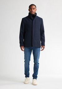 Marineblaue Wollmischungskapuze mit hohem Kragen, Druckknöpfen und seitlichen Taschen, kombiniert mit hellblauen Jeans und cremigen Sneakers.