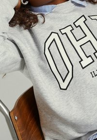 Grå sweatshirt med fet svart text "OH" och "ILL" på framsidan, lagd över en blå och vit randig skjorta med krage.