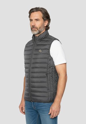 Grijze geïsoleerde bodywarmer met horizontaal doorgestikt patroon, hoge kraag en ritssluiting aan de voorkant; logopatch op de borst; gedragen over een wit shirt.