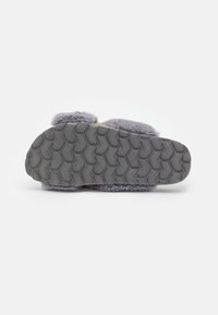 Chaussons en peluche gris avec une semelle en caoutchouc texturée présentant un motif en arêtes de poisson. L'intérieur est doux, tandis que l'extérieur a une finition duveteuse.