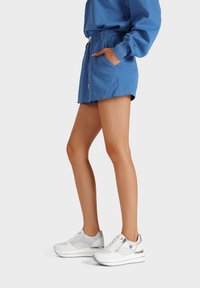 Shorts sportivi blu con vita elastica e tasche, abbinati a sneaker bianche e argentate con dettagli in rete e una suola spessa.