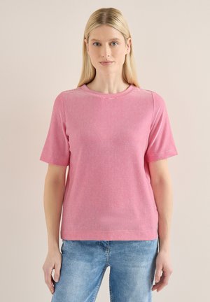 Femme aux cheveux blonds raides portant un haut rayé rose et blanc à manches courtes et un jean bleu, sur un fond uni.