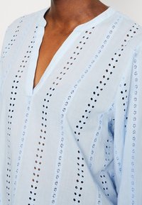 Blusa in cotone azzurro chiaro con scollatura a V, inserti in pizzo e dettagli ricamati con motivi a fenditura verticale.