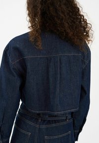 Tamna traper jakna skraćena i dugih rukava, s vidljivim šavovima i detaljem na leđima u obliku nabora. Glatka tekstura, klasičan dizajn.