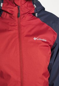 Röd och marinblå vindjacka tillverkad av slätt, lätt material; har en framzipper och justerbar huva. Columbia-logotyp synlig.