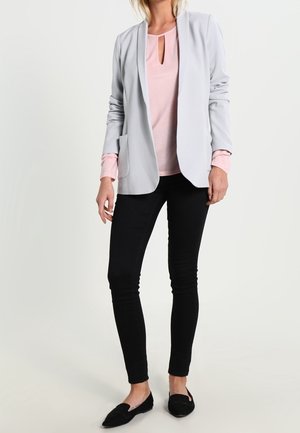 Vrouw die een lichtgrijze blazer draagt over een roze top, zwarte smalle broek en zwarte platte schoenen, staand tegen een effen achtergrond.