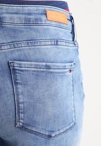 Jeans de mezclilla azul claro con un bolsillo trasero que presenta un pequeño acento rojo, superficie texturizada y una etiqueta de marca de cuero marrón.