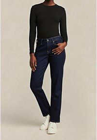 Personne portant un haut noir à manches longues, un jean bleu foncé et des chaussures blanches, debout avec une main dans la poche sur un fond uni.