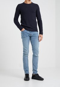 Man med marinblå långärmad skjorta, ljusblå tajta jeans och svarta sneakers, står med en hand i fickan mot vit bakgrund.