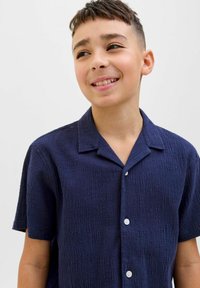 Námornícka košeľa s krátkym rukávom z textúrovanej látky, s kempovým golierom a predným zapínaním na gombíky. Hladké gombíky na léze.