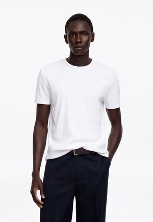 Homme portant un t-shirt blanc uni légèrement rentré dans un pantalon bleu marine foncé avec une ceinture noire tissée, debout devant un fond blanc.