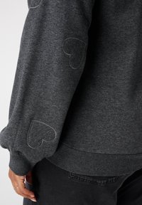 Donkergrijze trui met twee gestikte hartpatches op de mouw, gecombineerd met zwarte broek, hand rustend nabij de heup.