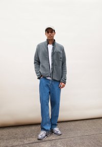 Giacca in pile grigia con zip e tasche laterali, abbinata a jeans blu e sneakers grigie. Lo sfondo è di un beige chiaro.