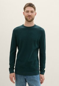 Suéter texturizado verde oscuro con cuello redondo, mangas largas y un pequeño logotipo en el dobladillo, combinado con jeans azul claro.