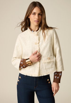 Vrouw draagt een crèmekleurig jasje met paillettenstructuur over een bruine blouse met polkadots, gecombineerd met donkerblauwe spijkerbroek met gouden knopen.