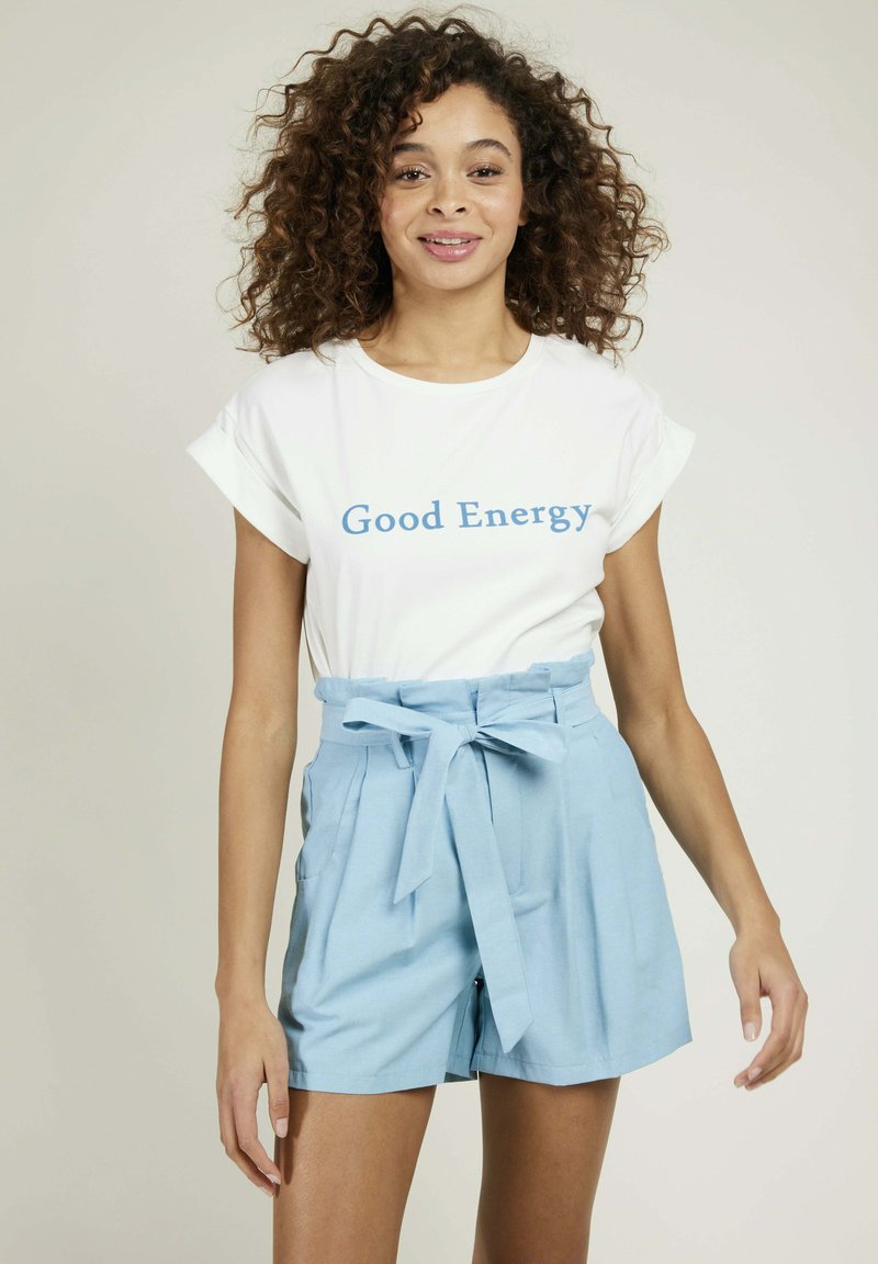 T-shirt blanc avec le texte bleu "Bonne Énergie", associé à un short taille haute bleu clair avec une ceinture nouée et des poches latérales.