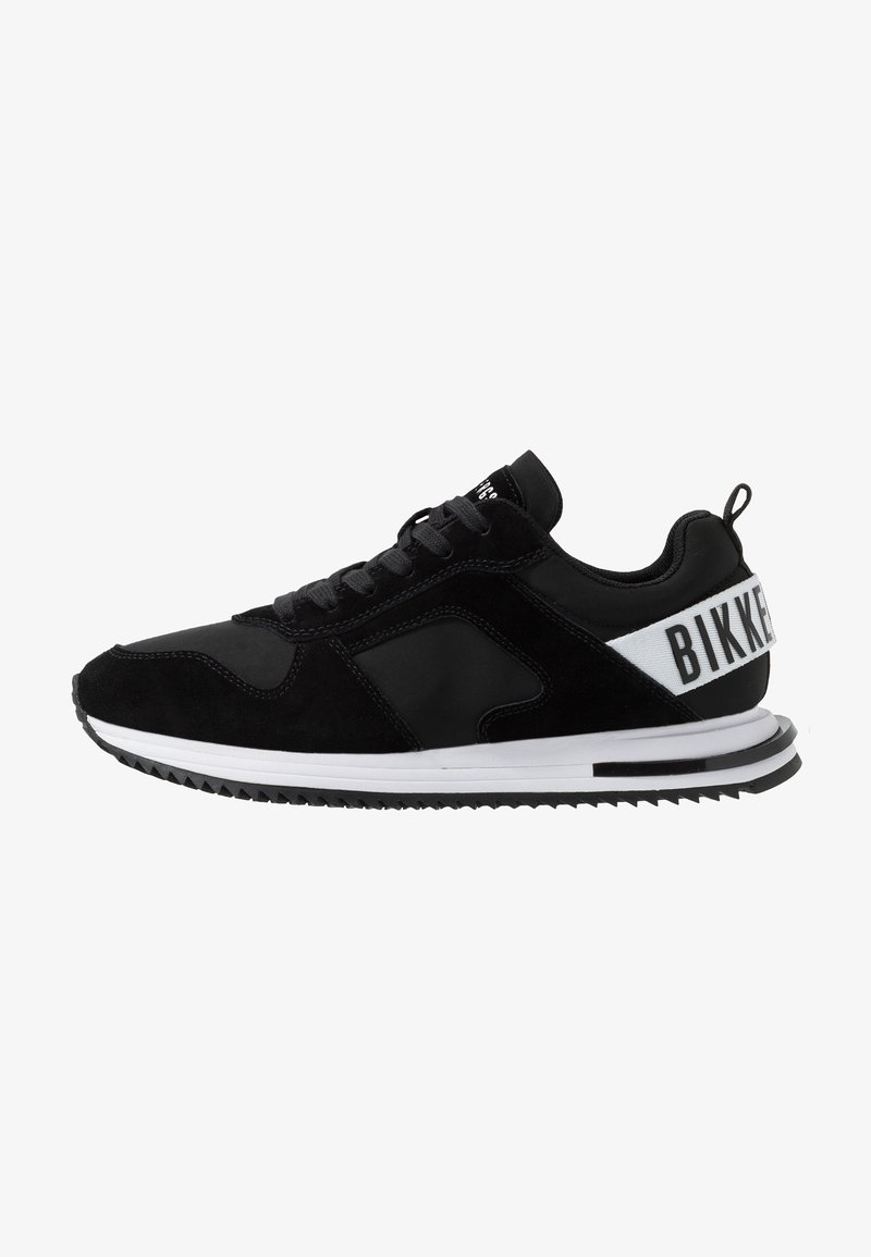 Baskets basses noires avec semelle blanche, lacets et bride de talon blanche avec texte noir "BIKK" sur le côté.