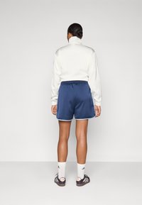 Chaqueta blanca con cremallera y cuello alto, combinada con pantalones cortos azul marino con ribete azul claro y una textura suave. Zapatillas deportivas blancas y negras.